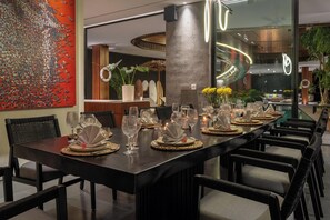 Een lange eettafel met meerdere stoelen, in een modern interieur met een groot abstract schilderij, potplanten en een ronde lamp.