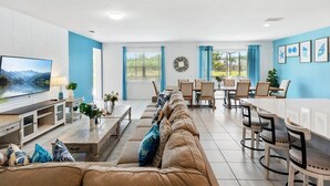 A spacious living room with a large flat-screen TV, a beige sofa, and a dining area with a white table and chairs.