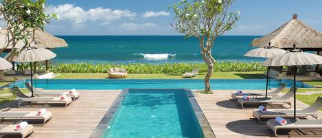 Zona de piscina con sillones de descanso y sombrillas, con vista a un océano azul cristalino.