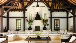Ein traditionell eingerichteter Raum mit Holzbalkendecke, einer zentralen Buddha-Statue, weißen Sofas und Zimmerpflanzen.