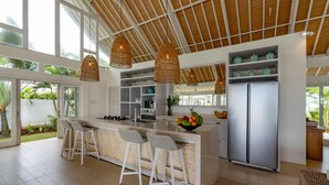 Una cocina moderna con una isla grande, taburetes de bar blancos, un refrigerador y lámparas colgantes de mimbre.