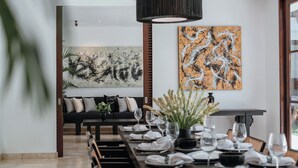 A dining area with a long table set for a meal, a black sofa with cushions, and abstract paintings on the walls. - Luxe Vacay Amara Pradi Chef and Staff included  lvid5 (Seminyak)