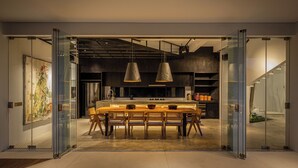Ein modernes Interieur mit einer Glastrennwand, einem zentralen Esstisch mit Stühlen, Pendelleuchten und einer Treppe im Hintergrund.