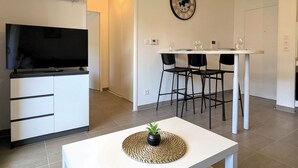 Una sala de estar moderna con una televisión de pantalla plana, una mesa de comedor con sillas, un reloj fijado a la pared y una pequeña planta en maceta sobre una mesa de centro.