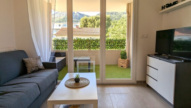 Un salón moderno con un sofá gris, una mesa de centro blanca, un televisor de pantalla plana sobre un mueble bajo y un ventanal corredero que da a un balcón con vistas a una montaña y vegetación.
