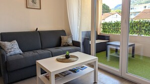 Una sala de estar moderna con un sofá gris, una mesa de centro blanca y un ventanal de vidrio que da a un balcón con vistas a casas y vegetación.