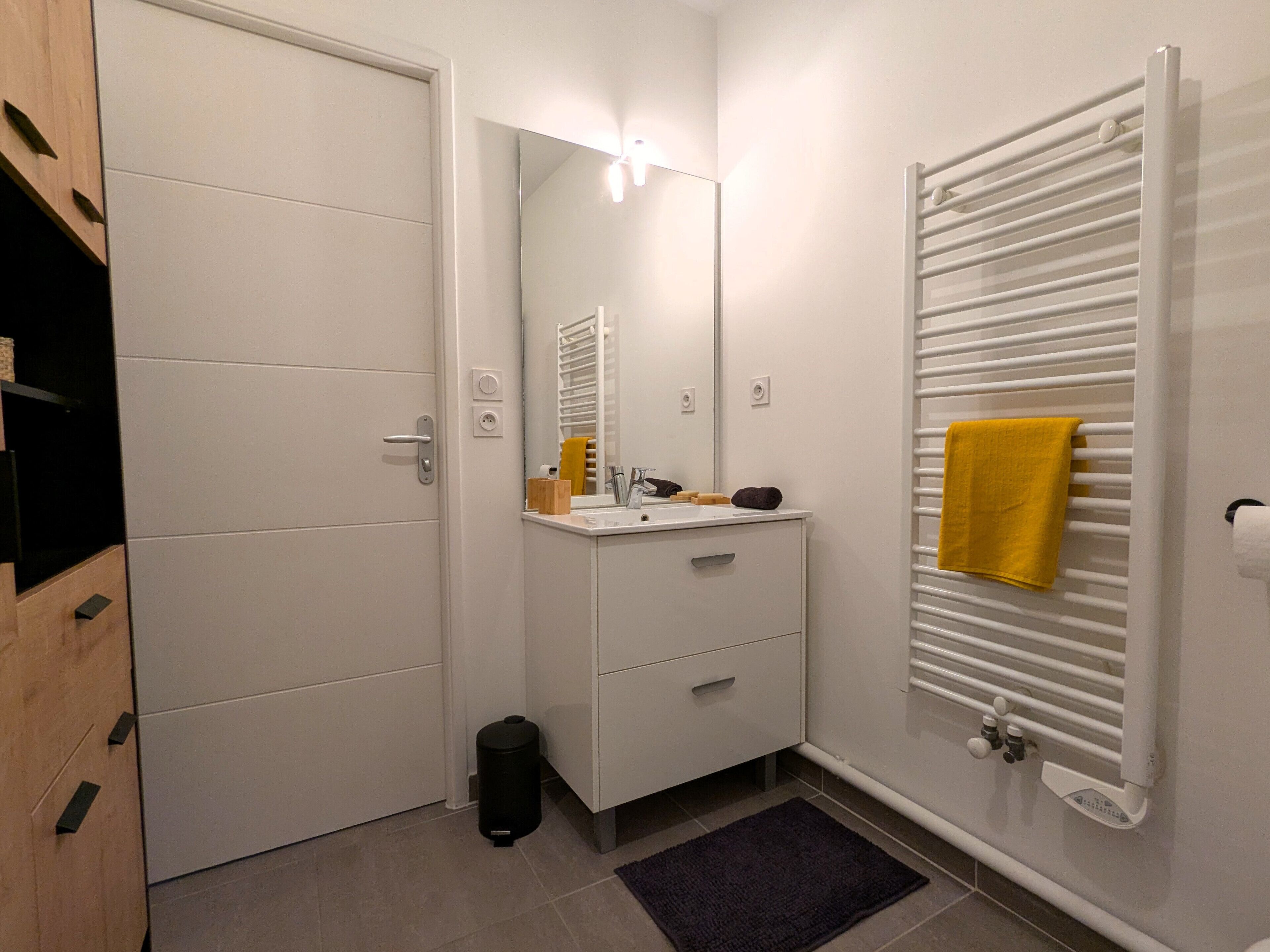 Un baño moderno con un tocador blanco, un espejo, un perchero con una toalla amarilla, un radiador y un cubo de basura negro.
