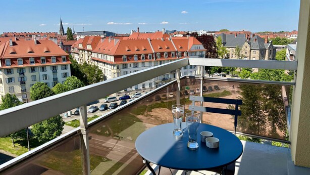 Un balcon avec une table et des chaises, donnant sur un paysage urbain composé d’immeubles aux toits rouges et d’une rivière.