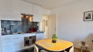Une cuisine compacte avec des meubles blancs, un évier en acier inoxydable, un four encastré et un lave-vaisselle de petite taille. On y trouve une petite table ronde avec deux chaises et une plante en pot.