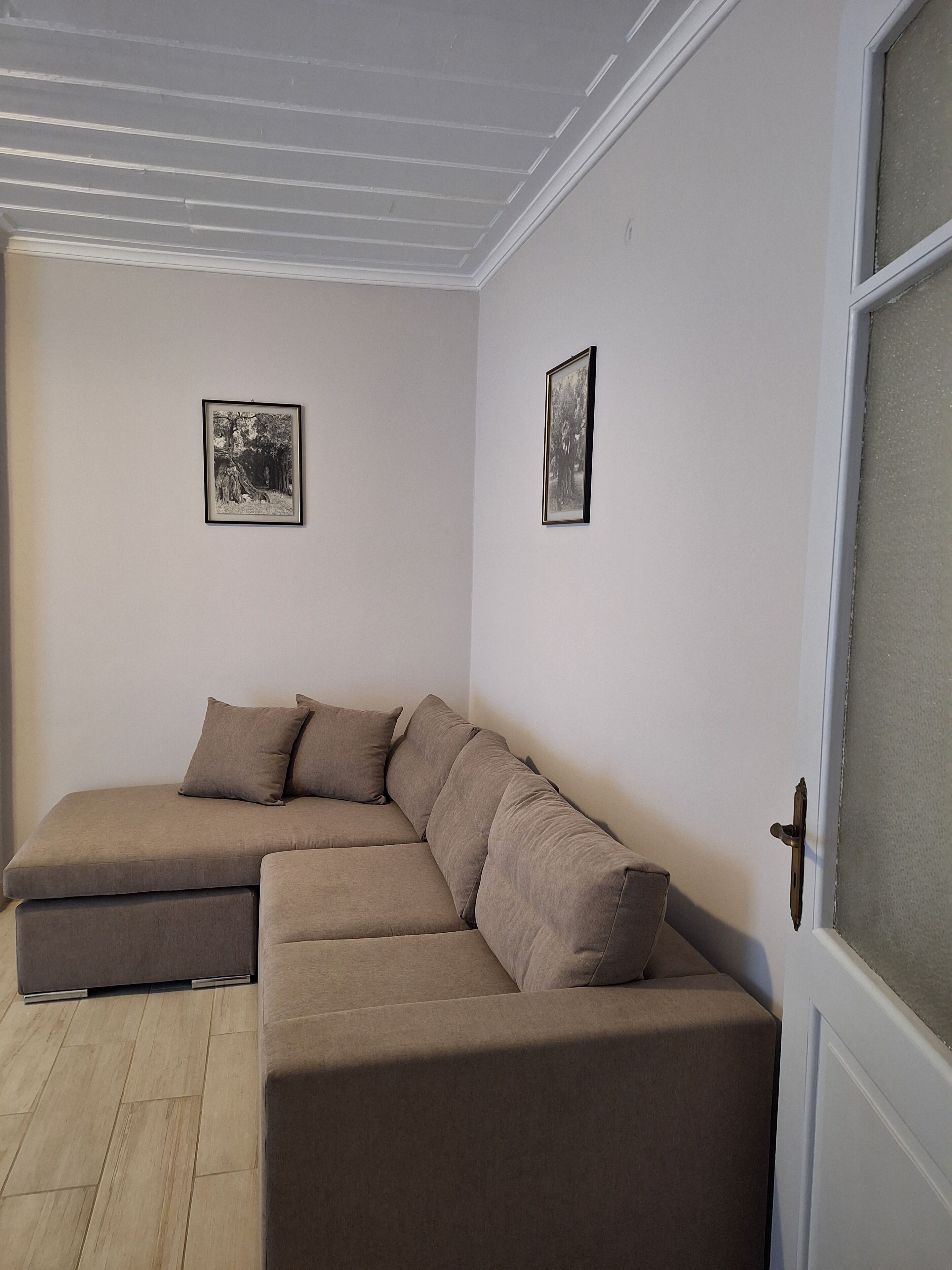 A corner living room with a beige sectional sofa, two framed pictures on the walls, and a white door with a frosted glass panel.