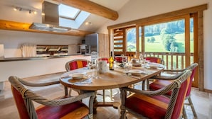 A dining area with a wooden table set for four, surrounded by wicker chairs with red cushions. A kitchen with modern appliances and a skylight is visible in the background. - Wifi, 2nd floor, balcony, fireplace or stove, tv, ski locker, 85m², Le Grand Bornand (Le Grand-Bornand)