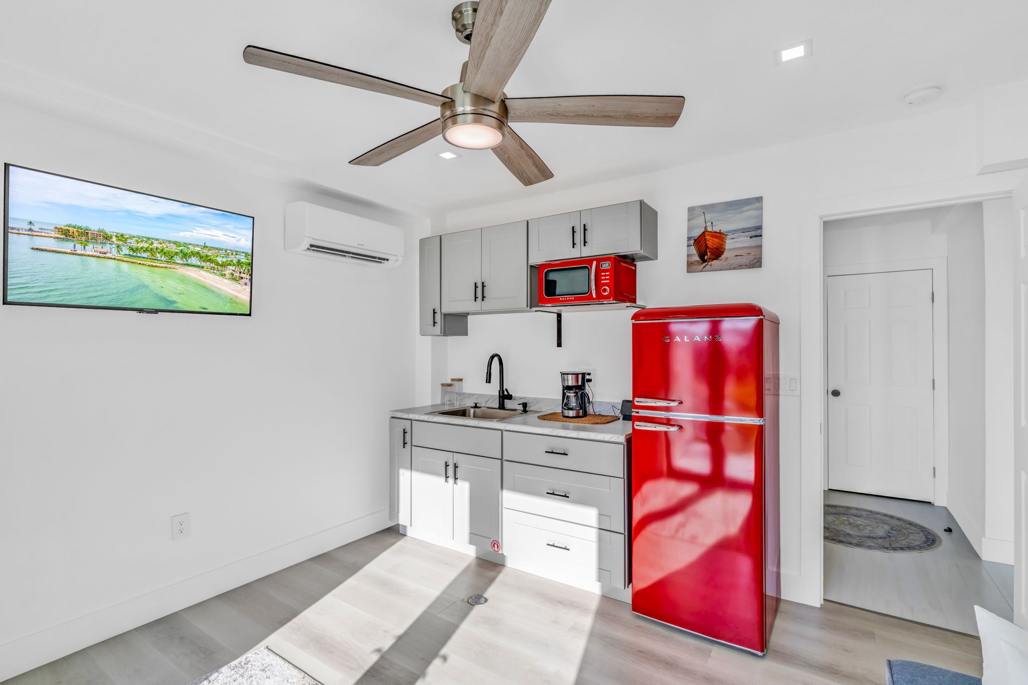 A modern kitchen with white cabinets, a red refrigerator, a ceiling fan, and a TV displaying a beach scene.