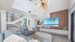 Een moderne woonkamer met een grijze bank, een poef en een flatscreen televisie aan een houten wand.