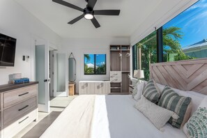 A modern bedroom with a bed, a ceiling fan, a wardrobe, and a large window with a view of palm trees.