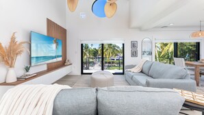 Een moderne woonkamer met een grote televisie, grijze banken, een plafondventilator en uitzicht op een zwembad en palmbomen.