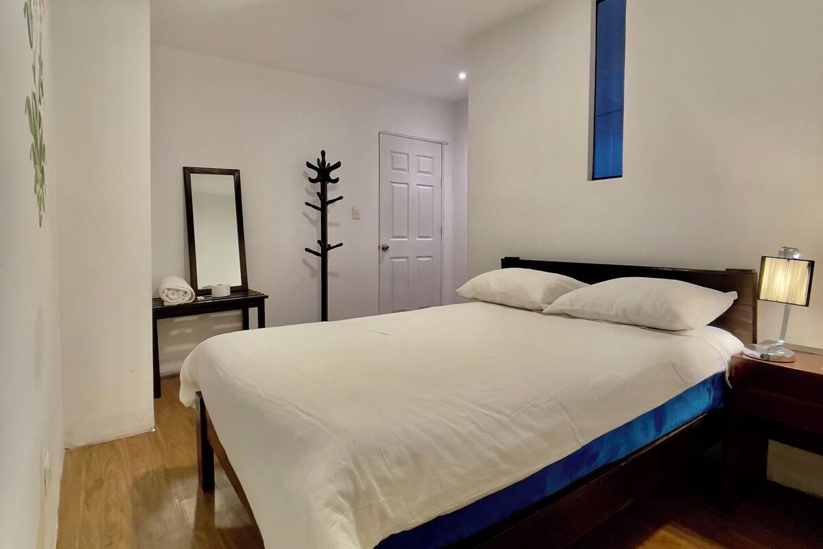 A bedroom with a large bed, a bedside table with a lamp, a mirror, and a door.