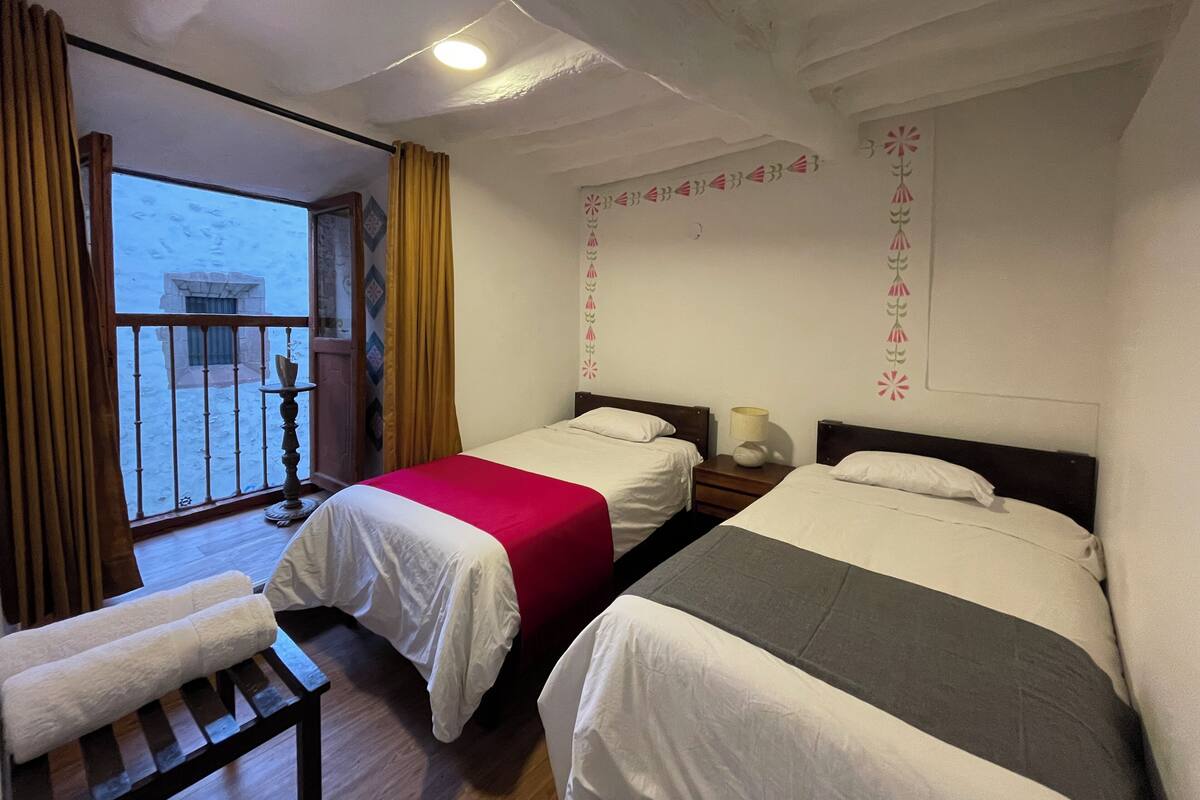A hotel room with two beds, a balcony with curtains, and a wooden bench.