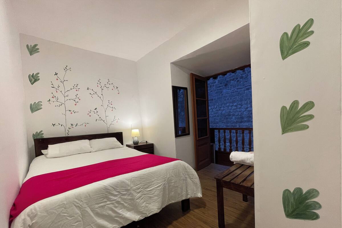A bedroom with a bed, a nightstand, a lamp, a wooden bench, and a balcony with a blue wall.