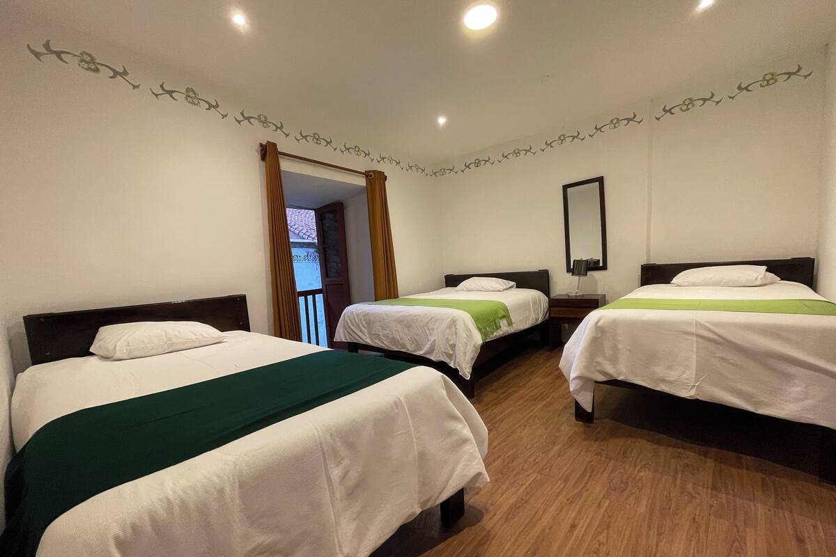 A hotel room with three single beds, wooden flooring, a small bedside table with a lamp, and a mirror on the wall.