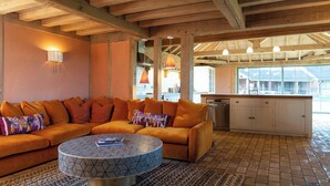 Ein modernes Wohnzimmer mit einem Ecksofa, einem Couchtisch mit Muster und einer Küchenzeile mit weißen Schränken und Pendelleuchten.