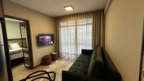 Una habitación de hotel con cama, sofá, televisión, ventana con cortinas y una mesita.