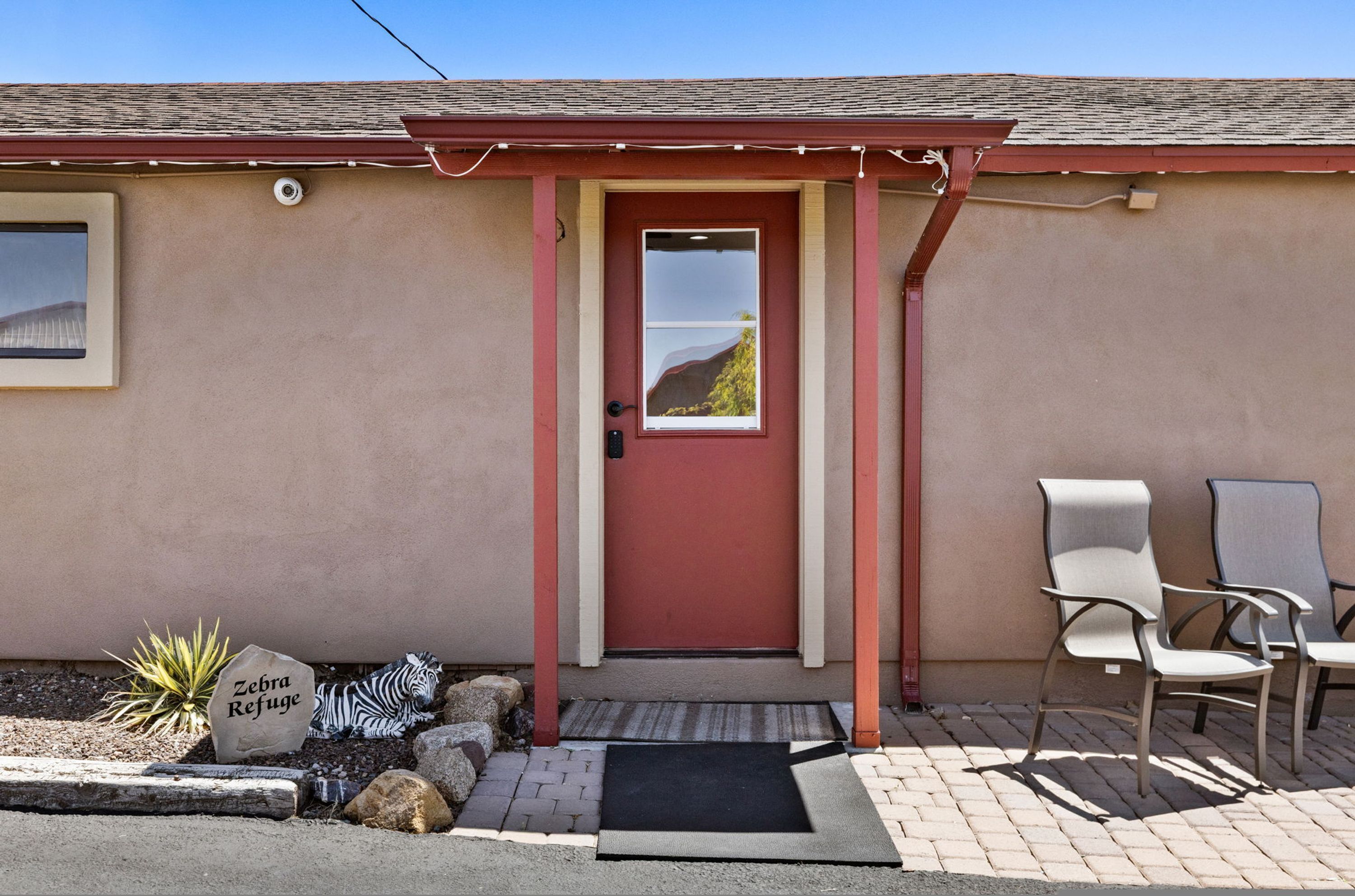 A building with a red door, a small window, and a sign reading "Zebra Refuge" on a rock. There are two chairs outside.