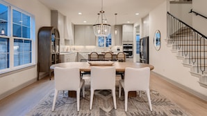 A modern kitchen with a dining area, featuring a chandelier, white chairs, and a staircase.