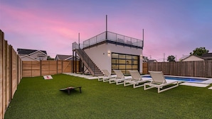 A backyard with artificial grass, a pool, and a modern house with a balcony.