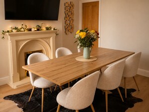 Un coin repas avec une table en bois, six chaises, un vase de fleurs, une cheminée et un téléviseur fixé au mur.
