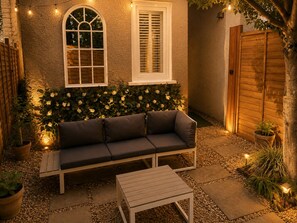 A cozy outdoor seating area with a sofa and table, surrounded by a garden with potted plants and a wooden fence.