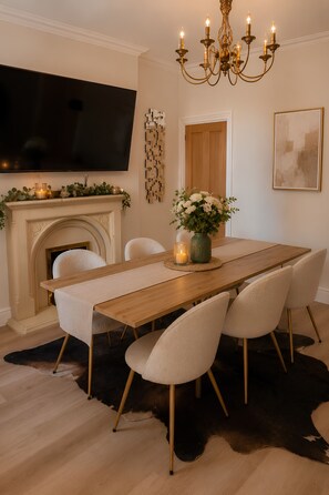A dining room with a wooden table, white chairs, a chandelier, a flat-screen TV, a fireplace, and a mounted artwork.