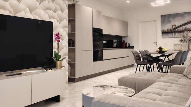 A modern living room with a flat-screen TV, a grey sofa, a dining area with a table and chairs, and a kitchen in the background.