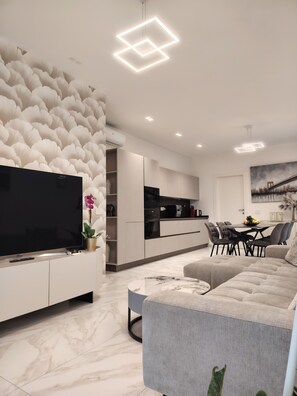 Un soggiorno moderno con una TV a schermo piatto, un divano grigio, un'area pranzo con tavolo e sedie, e una cucina sullo sfondo.