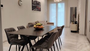 A modern dining area with a marble floor, a long table with chairs, a fruit bowl, and a cityscape painting on the wall.