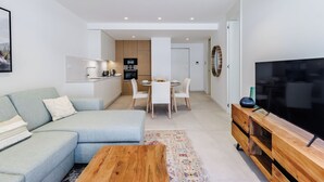 A modern living room with a sofa, coffee table, and TV stand. There is a dining area with a table and chairs. The kitchen is visible in the background with cabinets and appliances. - Santa Maria II Pool View (SR 1-B2.0O) (Lagos)
