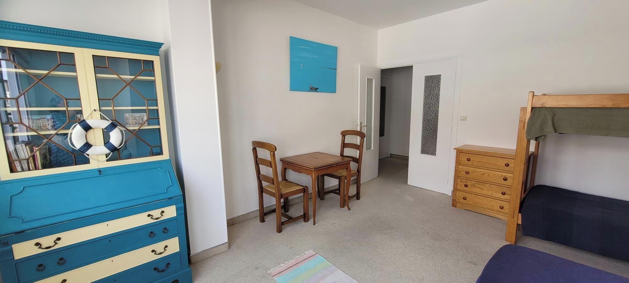 A room with a blue dresser, a wooden table with chairs, a bunk bed, and a door with a decorative panel.