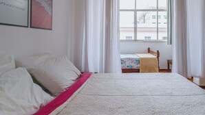 A bedroom with a bed, a bedside table, a window with curtains, and a view of buildings outside. - Apto Confort vel Tima Localiza o e pet Friendly (Teresópolis)