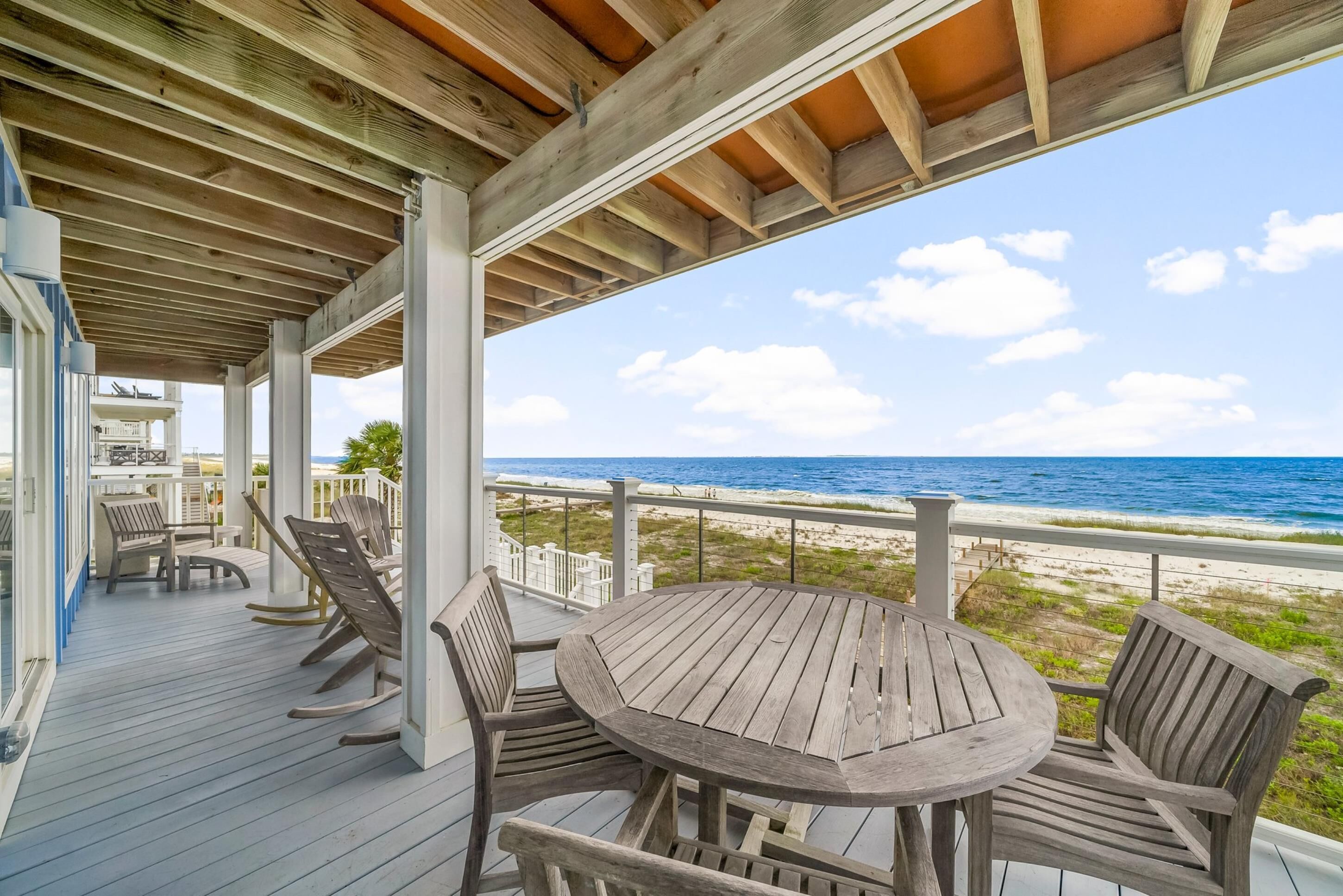 A wooden deck with a round table and chairs, overlooking a beach and ocean.