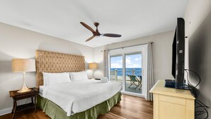 Habitación de hotel con una cama grande, ventilador de techo, televisión y vista al océano a través de una puerta corrediza de vidrio.