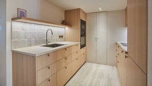Una cocina moderna con armarios de madera clara, encimera blanca, pileta de acero inoxidable y horno empotrado.
