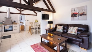 Sala de estar con un sofá de cuero marrón, una mesa de centro de madera y una alfombra roja. Se ve una cocina al fondo con armarios blancos y piso de baldosas.
