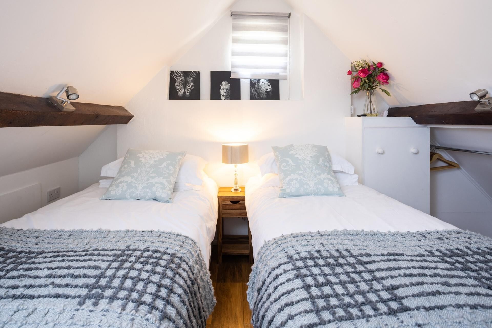 A bedroom with two beds, a nightstand, a lamp, a window with blinds, and a small cabinet.