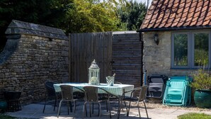 An outdoor patio with a stone wall, a wooden fence, a table with chairs, a lantern, and a window overlooking a garden. - The Dovecote 3-bed Cottage, Rowden Manor (Chippenham)