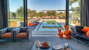 Een moderne woonkamer met uitzicht op een zwembad, grijze banken met oranje kussens, en een eethoek met een tafel gedekt voor twee personen.