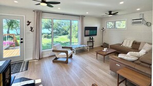 A spacious living room with a large sliding glass door leading to a garden, a ceiling fan, a flat-screen TV, and a brown leather sofa. - BEACHFRONT COTTAGE *fully renovated* (Saint Laurent)