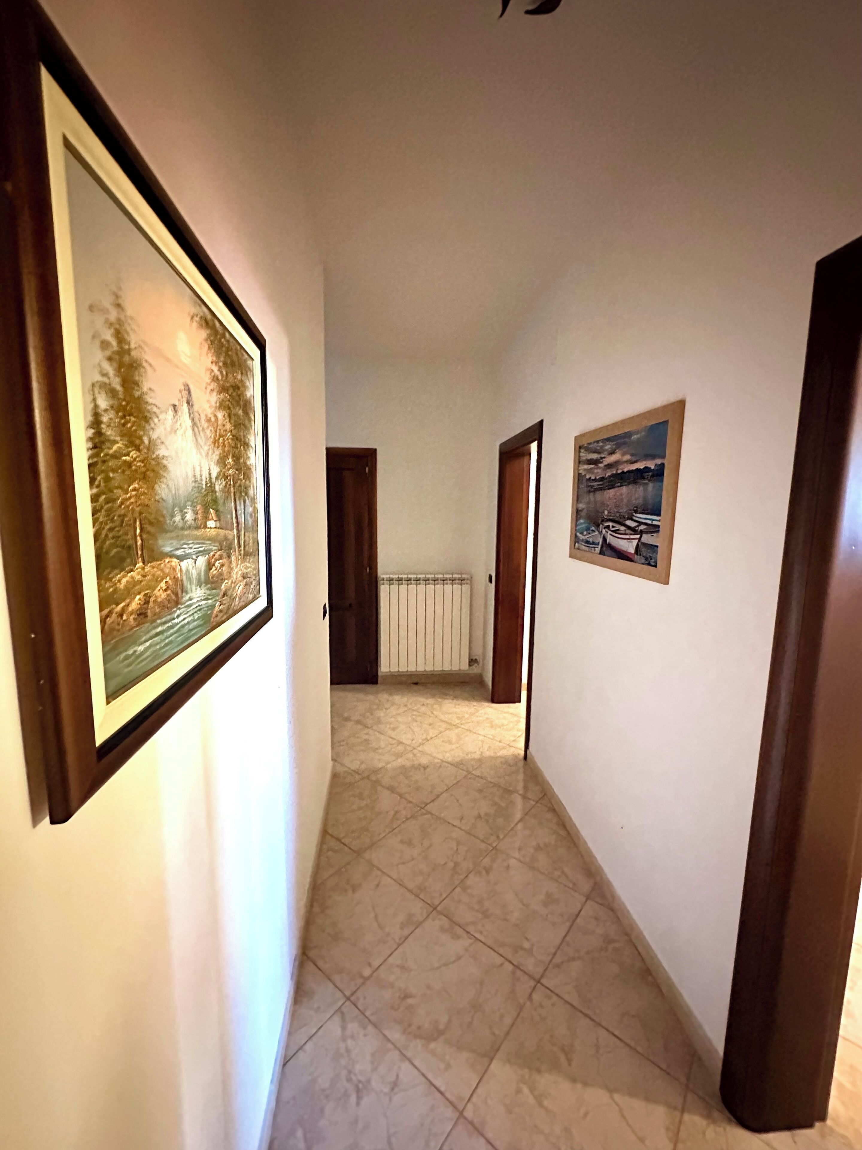 Um corredor com piso de azulejos, um quadro de paisagem à esquerda e um quadro de paisagem marítima à direita.
