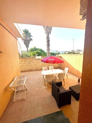 Uma área de pátio com mesa e cadeiras, um guarda-sol vermelho e uma pequena mesa branca com estrutura metálica.