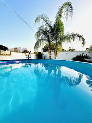 Piscina azul clara com escada, rodeada por um muro branco e palmeiras.
