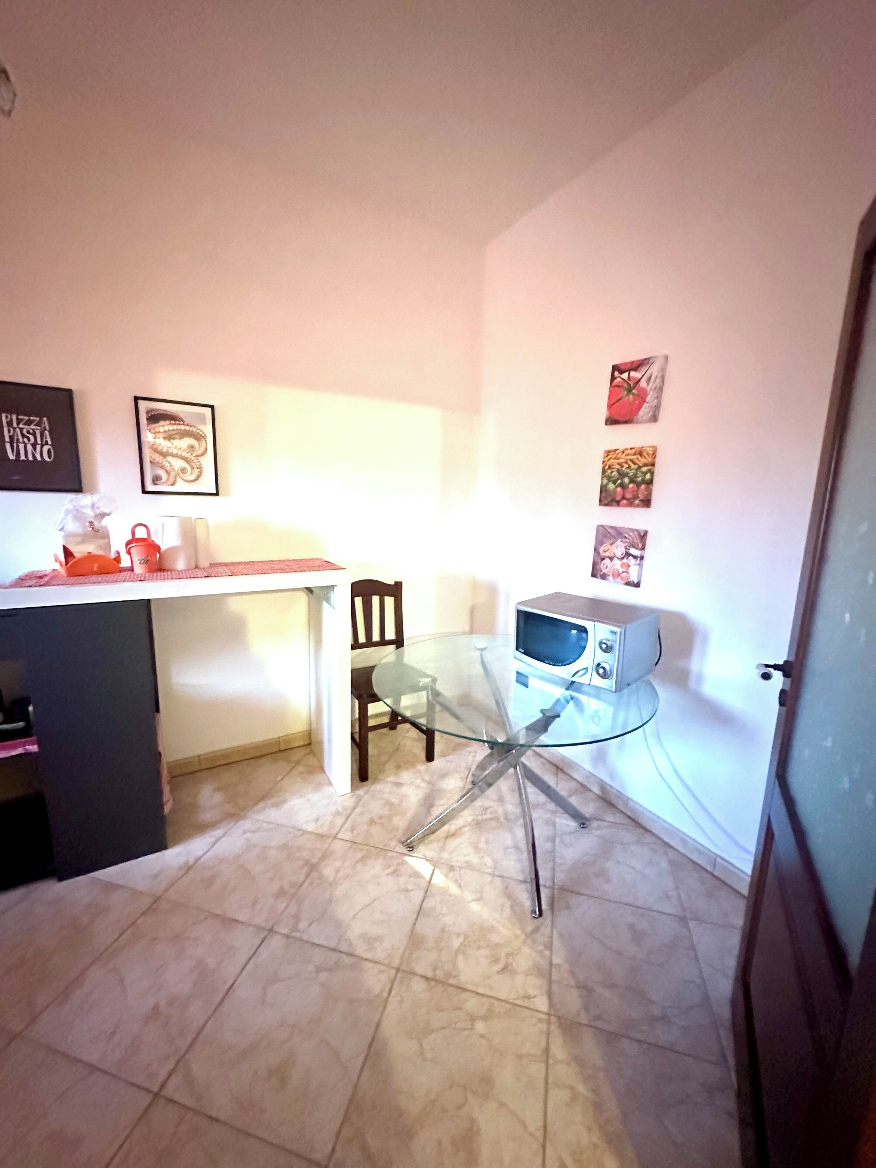 Uma cozinha pequena com uma mesa de vidro, micro-ondas e uma cadeira. Há quadros nas paredes e um letreiro de pizza.