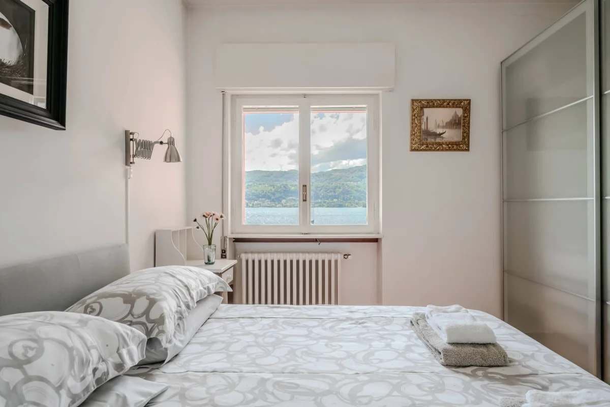 A bedroom with a bed, a radiator, a window with a view, a bedside lamp, and a framed picture on the wall.
